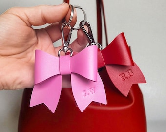May include: Two leather bow keychains with silver-toned metal lobster clasps and split rings. One bow is light pink with the embossed initials J.W, and the other is red with the embossed initials RB. Both are positioned in front of a red handbag.
