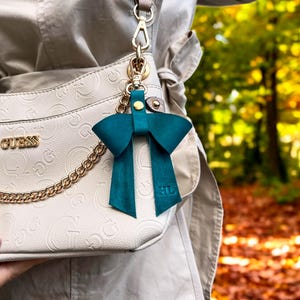 May include: A teal leather bow bag charm featuring a gold-tone metal clasp and the embossed initials HL on the bottom right tail. The charm is attached to a beige Guess handbag with a gold chain detail and a matching beige shoulder strap.
