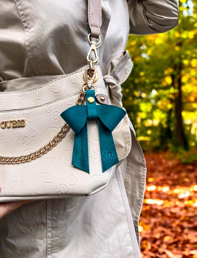 May include: A teal leather bow bag charm featuring a gold-tone metal clasp and the embossed initials HL on the bottom right tail. The charm is attached to a beige Guess handbag with a gold chain detail and a matching beige shoulder strap.