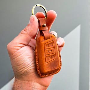 May include: A tan leather car key fob with a brass-coloured key ring. The fob features three embossed button icons and a stitched border. The leather is a warm, medium brown colour.