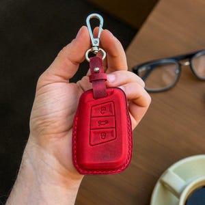 May include: A red leather car key fob cover with a silver-colored clasp. The fob cover has three embossed key symbols and a matching leather strap. The background is blurred, with a cup of coffee and eyeglasses visible.