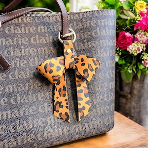 Puede incluir: Un bolso tote de cuero marrón con un llavero de lazo de estampado de leopardo. El bolso tiene un asa de cuero marrón y un cierre de cremallera. El llavero está unido a un anillo de metal dorado.