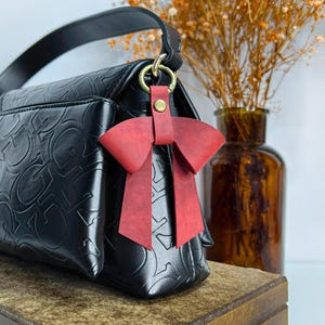 May include: A black handbag featuring an embossed repeating letter pattern and a single top handle. A red leather bow charm is attached to the bag's gold-toned metal ring hardware with a small brass-colored snap button fastener.