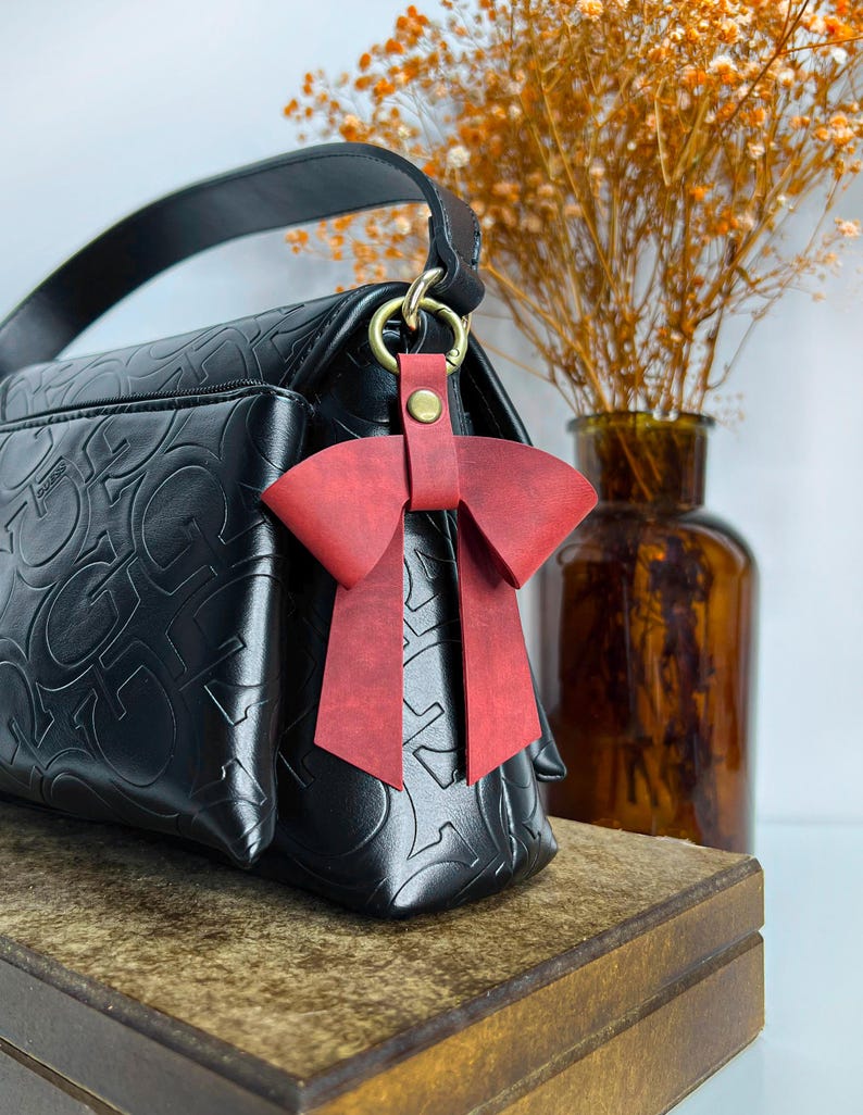 May include: A black handbag featuring an embossed repeating letter pattern and a single top handle. A red leather bow charm is attached to the bag's gold-toned metal ring hardware with a small brass-colored snap button fastener.
