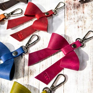 May include: A collection of leather bow keychains in red, magenta, blue, yellow, and brown, each featuring a silver metal lobster clasp, a central silver rivet, and the embossed letters ED on one of the bow tails, arranged on a white surface.