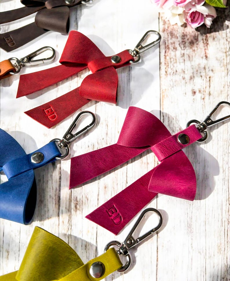 May include: A collection of leather bow keychains in red, magenta, blue, yellow, and brown, each featuring a silver metal lobster clasp, a central silver rivet, and the embossed letters ED on one of the bow tails, arranged on a white surface.