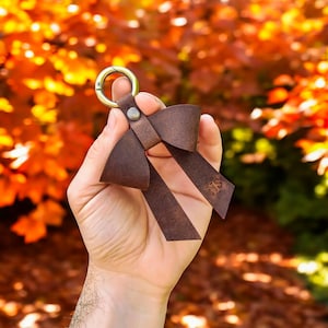 May include: A dark brown leather keychain shaped like a bow, featuring a brass-toned metal lobster clasp attached with a small rivet. The leather ribbon on the right side is embossed with the letters NS, creating a simple and functional accessory.
