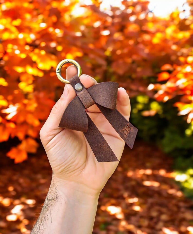 May include: A dark brown leather keychain shaped like a bow, featuring a brass-toned metal lobster clasp attached with a small rivet. The leather ribbon on the right side is embossed with the letters NS, creating a simple and functional accessory.