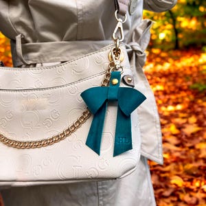 May include: A dark teal leather bow bag charm featuring a gold-tone metal snap closure and a gold chain attachment. The bow has two long tails with the embossed letters HL at the bottom, clipped onto a cream-colored handbag with a repeating logo pattern.