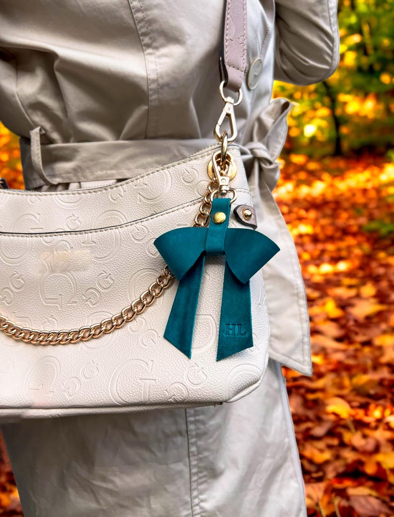 May include: A dark teal leather bow bag charm featuring a gold-tone metal snap closure and a gold chain attachment. The bow has two long tails with the embossed letters HL at the bottom, clipped onto a cream-colored handbag with a repeating logo pattern.