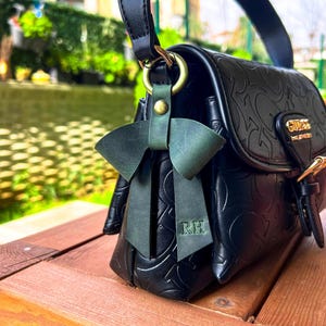 May include: A black handbag with an embossed logo pattern and a gold-tone metal clasp featuring the word GUESS. A dark green leather bow charm is attached to the side ring, with the initials RH embossed on one of the hanging ribbon tails.