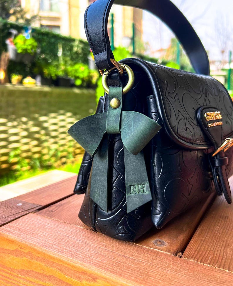May include: A black handbag with an embossed logo pattern and a gold-tone metal clasp featuring the word GUESS. A dark green leather bow charm is attached to the side ring, with the initials RH embossed on one of the hanging ribbon tails.