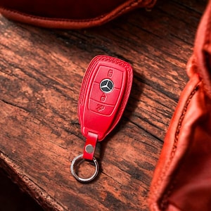 May include: A red leather key fob with a silver key ring. The key fob has a Mercedes-Benz logo and four buttons.