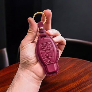 Op de afbeelding: Bordeauxkleurige leren sleutelhanger met een gouden ring en een sleutelgatontwerp. De sleutelhanger heeft vier knoppen met symbolen voor ontgrendelen, vergrendelen, kofferbak en paniek.