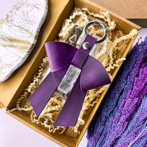 May include: A deep purple leather bow keychain featuring a silver-toned metal ring and a central snap fastener. The bow is attached to a clear, rectangular acrylic block, presented inside a brown cardboard box with shredded paper filler.