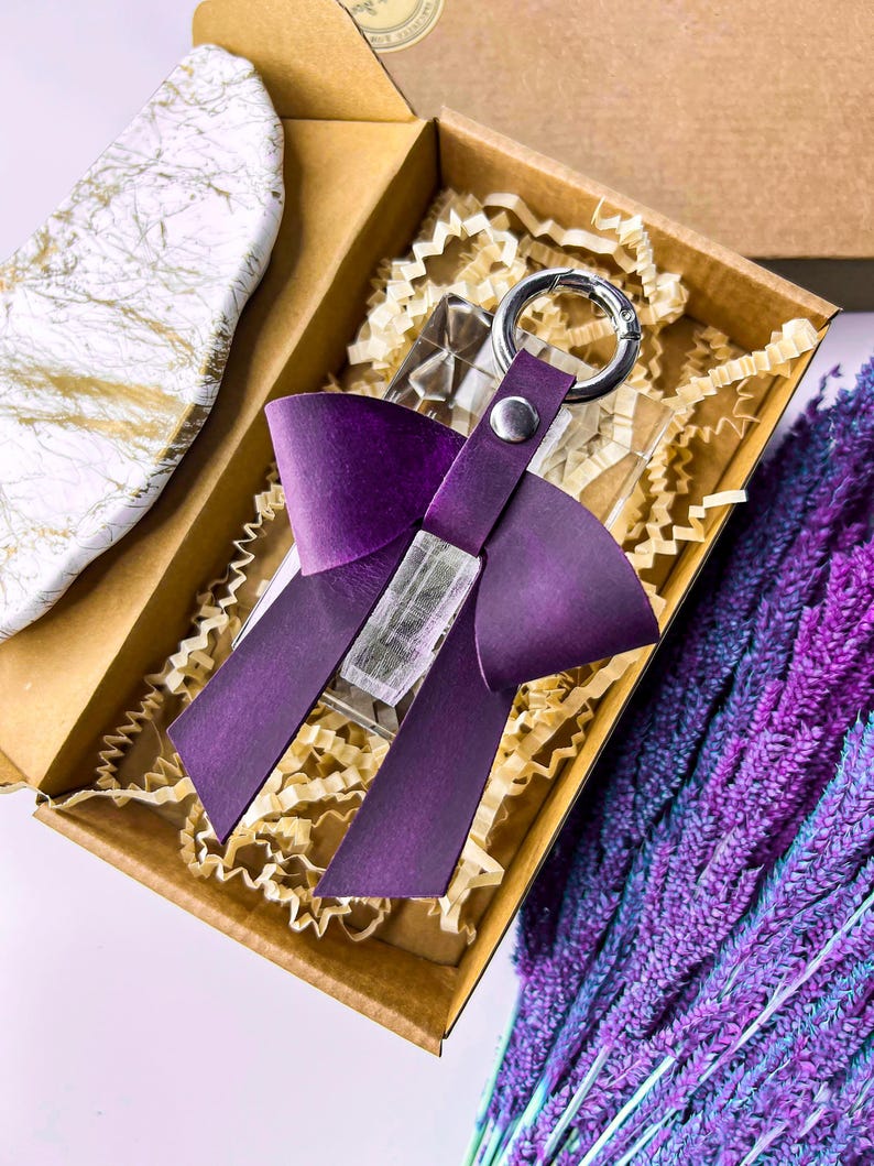 May include: A deep purple leather bow keychain featuring a silver-toned metal ring and a central snap fastener. The bow is attached to a clear, rectangular acrylic block, presented inside a brown cardboard box with shredded paper filler.