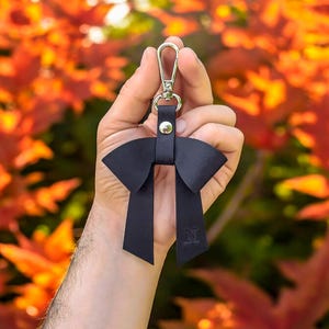 May include: A dark navy blue leather bow keychain featuring a silver-toned metal lobster clasp and a central silver rivet. The leather bow has two long, dangling tails, with the letter N embossed on the lower right tail of the accessory.
