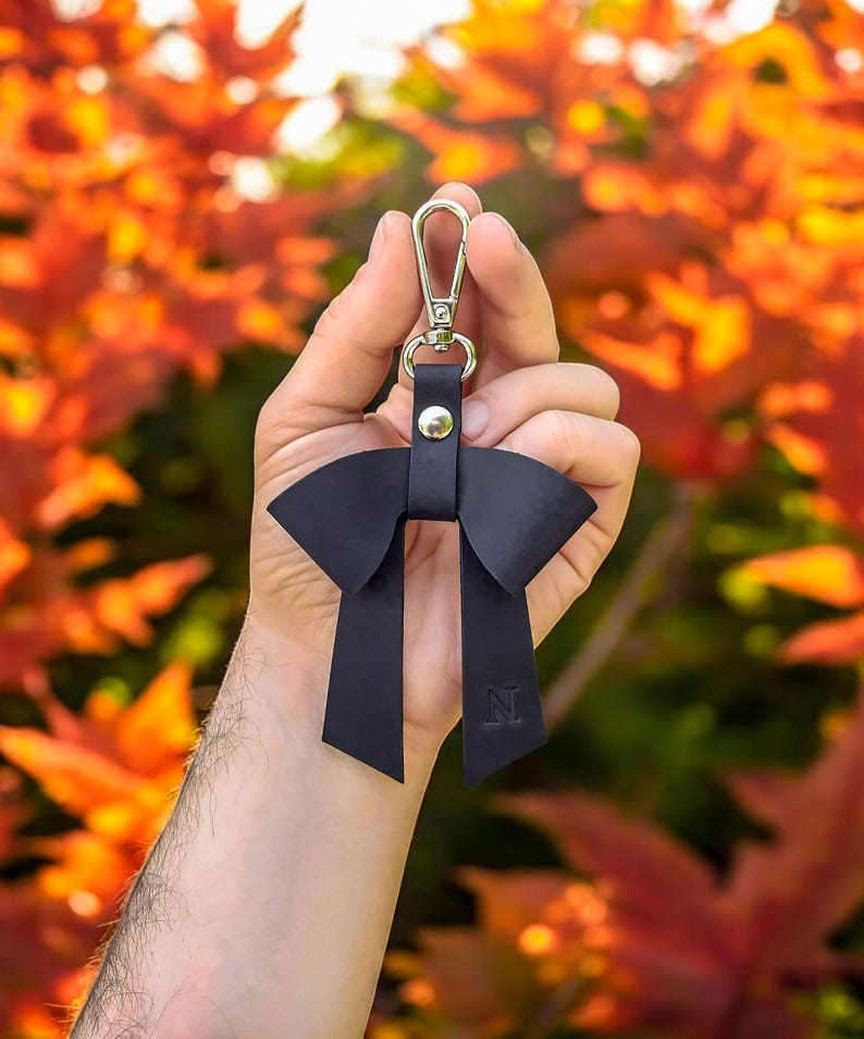 May include: A dark navy blue leather bow keychain featuring a silver-toned metal lobster clasp and a central silver rivet. The leather bow has two long, dangling tails, with the letter N embossed on the lower right tail of the accessory.
