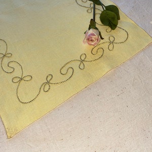 May include: A yellow linen placemat with a gold embroidered border design. A single pink rose with green leaves is placed on the mat.
