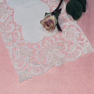 May include: White lace doily with a delicate floral pattern, featuring a single pink rose on a pink background.