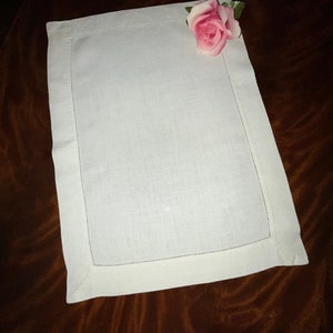 May include: A rectangular white linen placemat with a stitched border. A pink rose with green leaves rests on the upper right corner. The placemat is displayed on a dark brown surface.