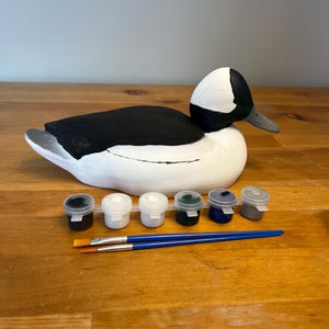 Puede incluir: Un señuelo de pato de madera blanco y negro con cabeza blanca y cuerpo negro. El señuelo está sentado sobre una superficie de madera con seis pequeños botes de pintura y dos pinceles.