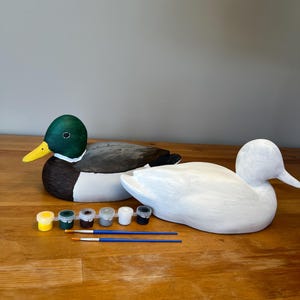 Puede incluir: Dos figuras de pato de madera, una pintada con colores naturales y la otra imprimada en blanco, están sobre una superficie de madera. Pequeños botes de pintura de varios colores y dos pinceles están colocados cerca, sugiriendo un proyecto de pintura DIY.