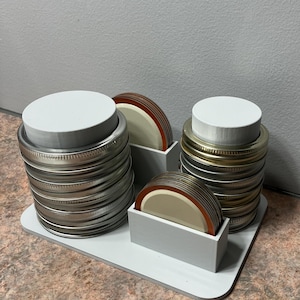 May include: White plastic organiser with three compartments for storing jar lids. The organiser has two stacks of metal jar lids and one stack of white and orange jar lids.
