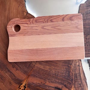 May include: A rectangular wooden cutting board with a hole in the top left corner. The board is made of light and dark wood, creating a striped pattern. The board is sitting on a wooden surface.