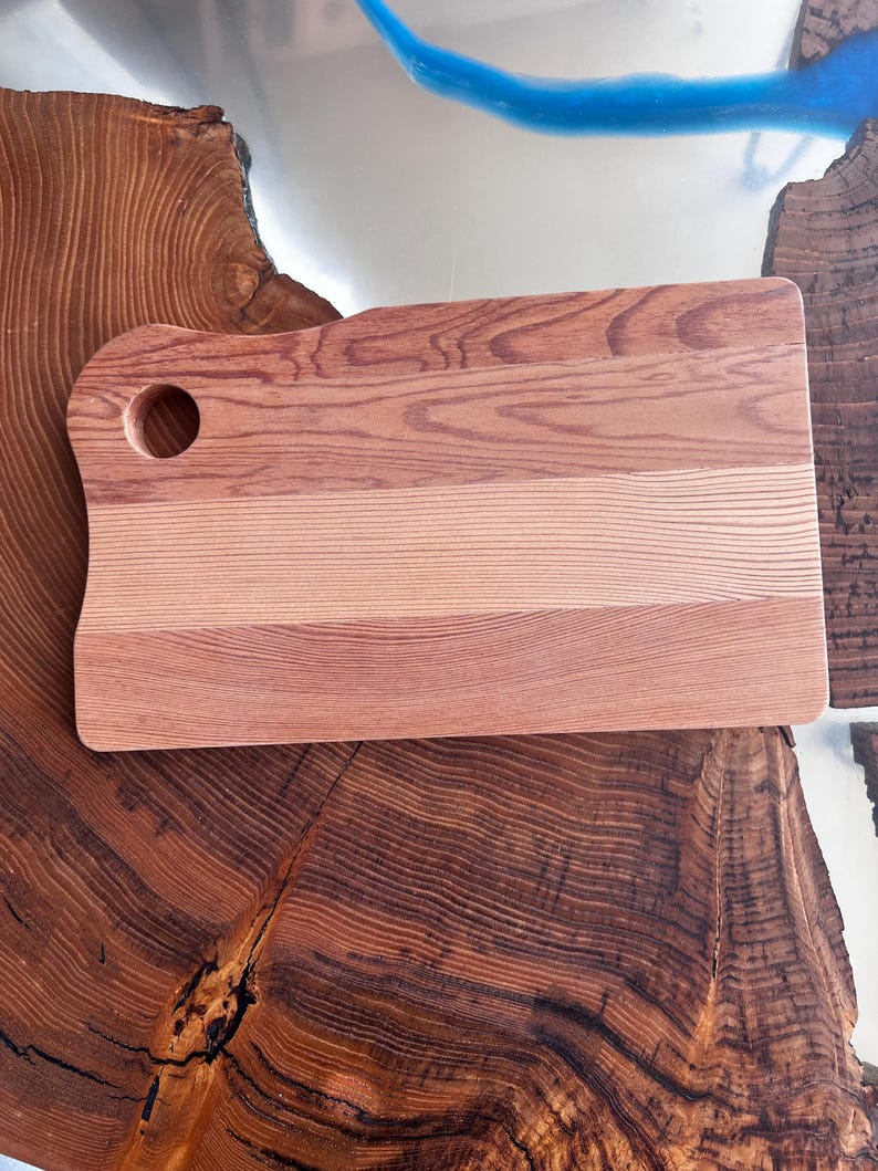 May include: A rectangular wooden cutting board with a hole in the top left corner. The board is made of light and dark wood, creating a striped pattern. The board is sitting on a wooden surface.