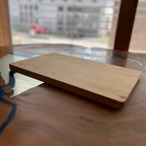 Op de afbeelding: Een lichtbruin houten snijplank met een glad oppervlak. De plank ligt op een houten tafel met een blauwe en bruine epoxyhars inlay.