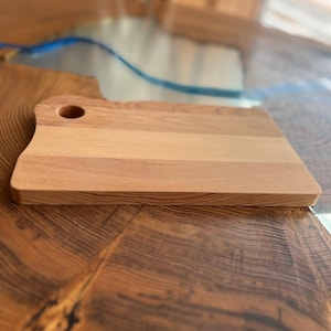 May include: A rectangular wooden cutting board with a small hole in the top. The board has a light brown color and a smooth surface.