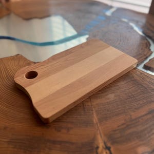 May include: A natural wood cutting board with a rounded handle and a hole for hanging. The board has a light brown color and a smooth surface.