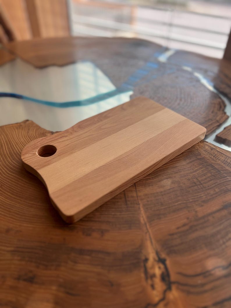 May include: A natural wood cutting board with a rounded handle and a hole for hanging. The board has a light brown color and a smooth surface.