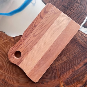 May include: A rectangular wooden cutting board with a handle and a hole in the handle. The board is made of two different types of wood, one light and one dark. The board is sitting on a wooden surface.