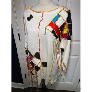 May include: A long-sleeve white top featuring a patchwork design with various coloured fabric squares in red, blue, yellow, purple, and black. The garment includes exposed seams with yellow and orange stitching and decorative hanging fabric fringe.