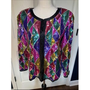 Puede incluir: Una chaqueta de manga larga con un vibrante patrón de rombos cubierto de lentejuelas multicolores en tonos morado, rosa, verde y dorado. La prenda tiene un borde negro sólido a lo largo del cuello redondo y un cierre frontal vertical de cuentas negras.