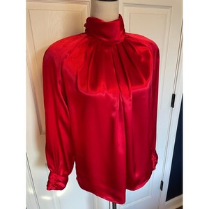 May include: A bright red satin blouse with a high, gathered neckline and long sleeves. The blouse has an elegant design with buttoned cuffs. This classic top is suitable for formal or business wear.