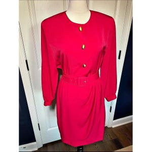 May include: A vibrant red long-sleeved dress featuring a round neckline, three gold-toned circular buttons down the center front, and a matching fabric belt with a rectangular buckle cinched at the waist, displayed on a mannequin.