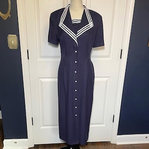 Puede incluir: Un vestido azul marino con cuello blanco y rayas blancas. El vestido tiene mangas cortas y una botonadura frontal. El vestido se exhibe en un maniquí.