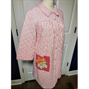 May include: A quilted housecoat featuring a red and white vertical stripe pattern, a collared neckline, and three-quarter length sleeves. The garment includes a front button closure and a single red pocket with white polka dots and a floral applique.
