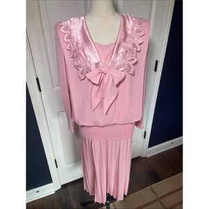 Può includere: Un abito rosa vintage con colletto in raso e fiocco. La parte superiore a maniche lunghe presenta un bordo in pizzo decorativo sul colletto e una gonna a pieghe. L'abito è esposto su un manichino.