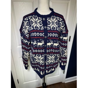 Könnte beinhalten: Marineblauer Cardigan-Pullover mit Schneeflocken-, Rentier- und geometrischem Muster in Weiß und Rot. Der Pullover hat einen runden Ausschnitt, lange Ärmel und eine Knopfleiste. Der Pullover wird auf einer Schaufensterpuppe präsentiert.