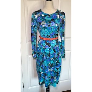 May include: A vintage floral print dress in shades of teal, blue, and purple. The long-sleeved dress features a button-down front, a pleated skirt, and a coral-coloured belt. The dress is displayed on a mannequin.