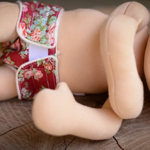 May include: A cloth doll with a floral diaper. The diaper is red with a white floral pattern and white velcro closures. The doll's body is a light beige color, and it is lying on a wooden surface.
