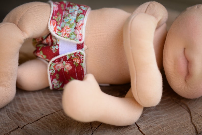 May include: A cloth doll with a floral diaper. The diaper is red with a white floral pattern and white velcro closures. The doll's body is a light beige color, and it is lying on a wooden surface.