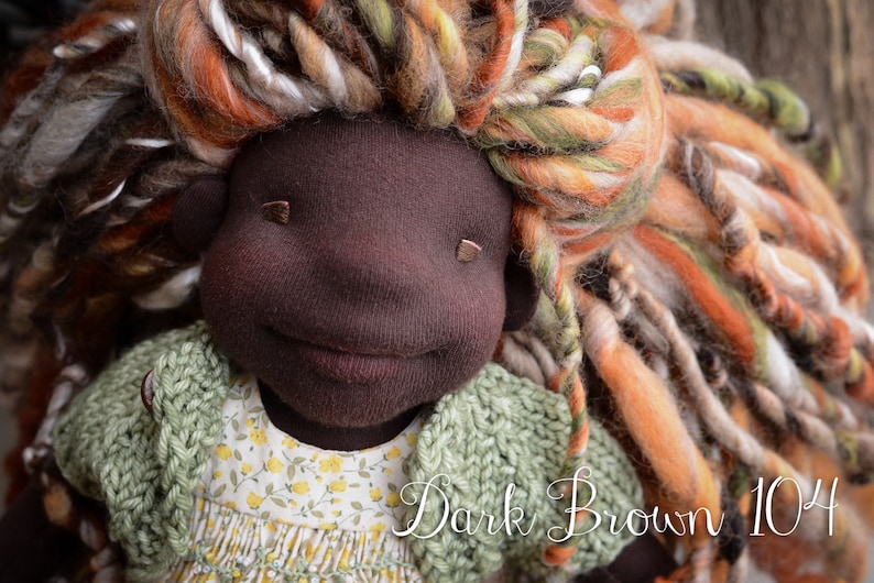 Op de afbeelding: Handgemaakte pop met een donkerbruine huid en een glimlach. De pop heeft lang, getextureerd haar in tinten oranje, bruin en wit. Ze draagt een groene gebreide trui en een bloemenjurk. De tekst "Dark Brown 104" is op de afbeelding te zien.