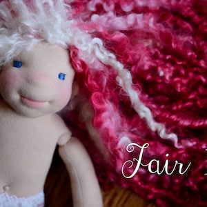 Op de afbeelding: Handgemaakte pop met een zacht stoffen lijfje en een lachend gezicht. De pop heeft blauwe ogen, roze wangen en wit en roze krullend haar. De tekst "Fair 115" staat op de afbeelding.