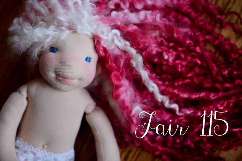 Op de afbeelding: Handgemaakte pop met een zacht stoffen lijfje en een lachend gezicht. De pop heeft blauwe ogen, roze wangen en wit en roze krullend haar. De tekst "Fair 115" staat op de afbeelding.