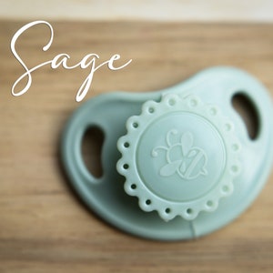 May include: A sage green silicone pacifier with a bee design on the front. The pacifier has a textured, scalloped edge.